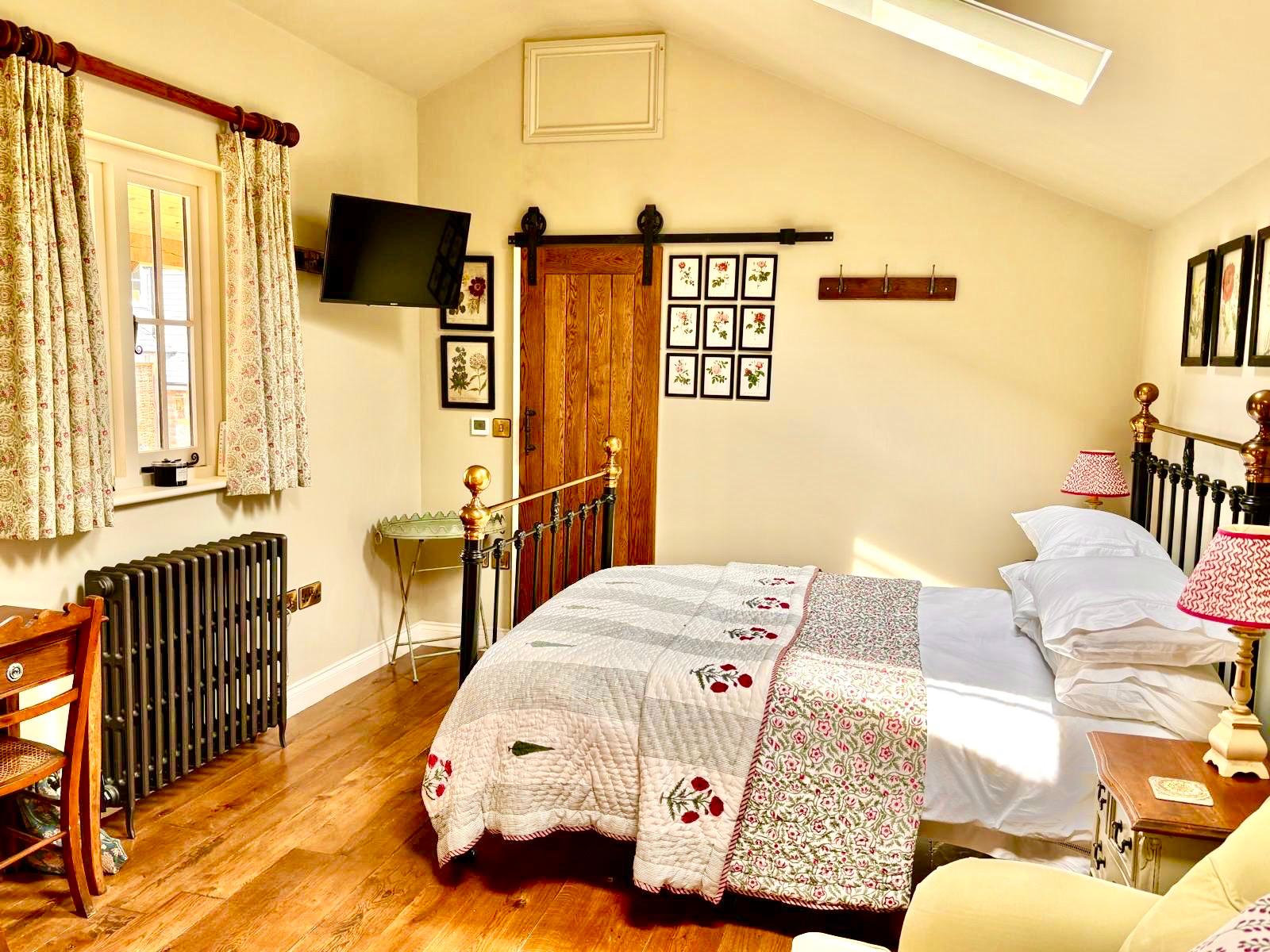 The room from the window side: a wall‑mounted TV, cast‑iron radiator, writing desk and the brass bed