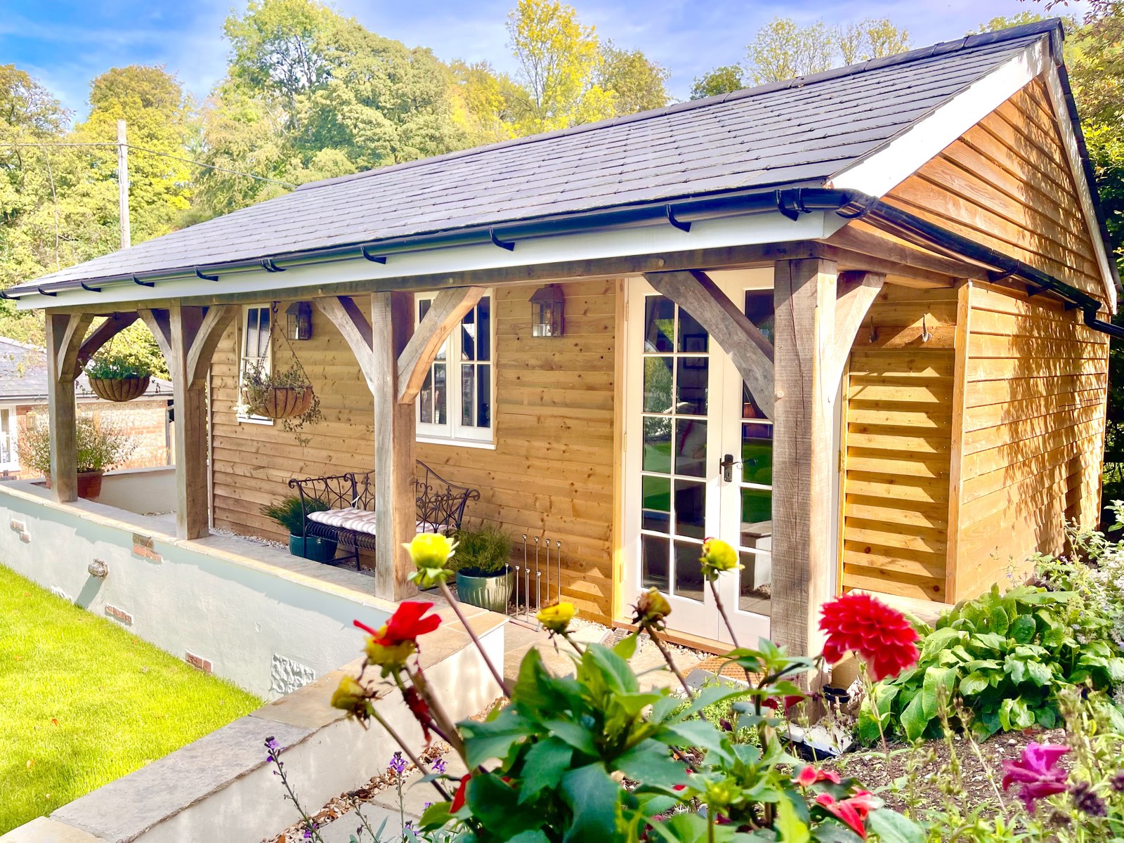 Garden Lodge: a larch‑clad cabin with an oak‑framed veranda and French doors, seen past red and yellow dahlias in the garden